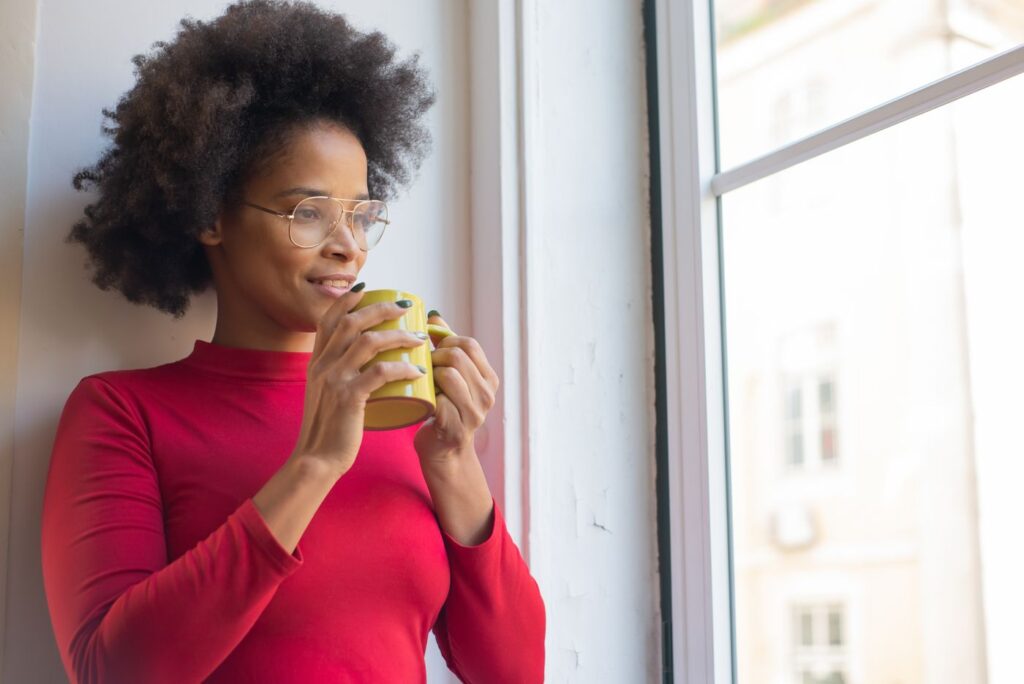 Woman in a red sweater smiling as she looks out a sunlit window, holding a cup